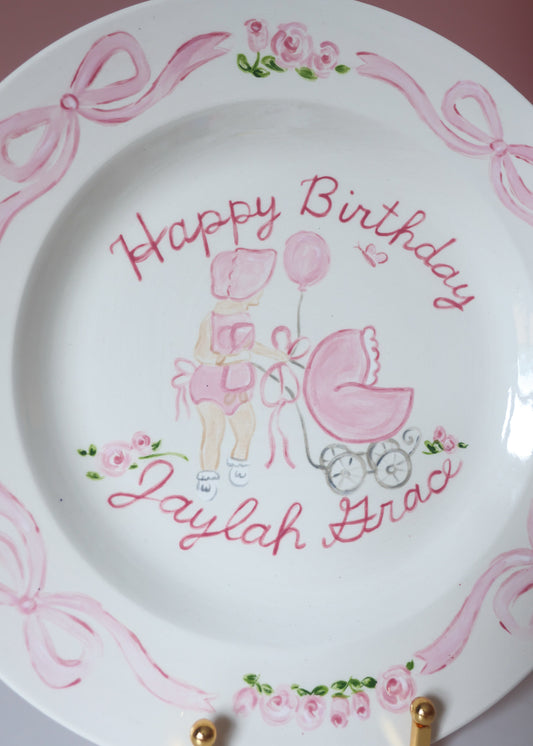 Decorative plate with 'Happy Birthday Jaylah Hadd' text and illustrations of a baby and stroller on a pink background.